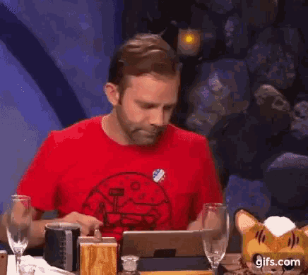 a man in a red shirt is sitting at a table with glasses of wine and a stuffed cat .