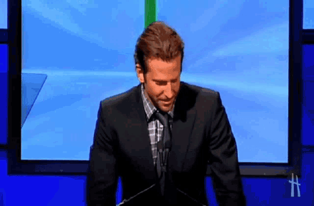 a man in a suit and tie is standing in front of a blue screen with the letter h on it