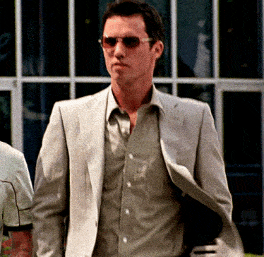 a man wearing sunglasses and a suit stands in front of a window