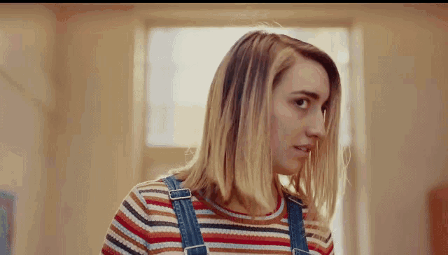 a woman wearing overalls and a striped shirt looks to the side