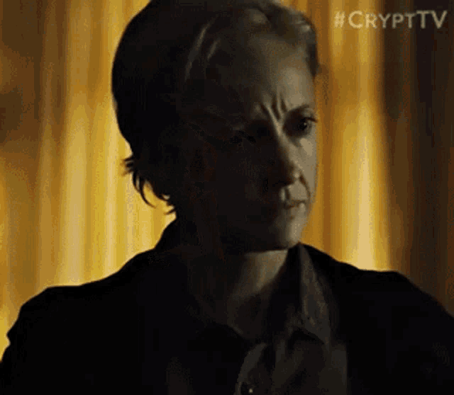 a woman in a dark room with a yellow curtain behind her is making a funny face .