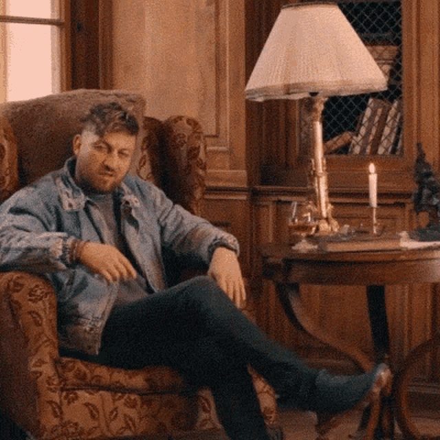 a man in a denim jacket sits in a chair with his legs crossed