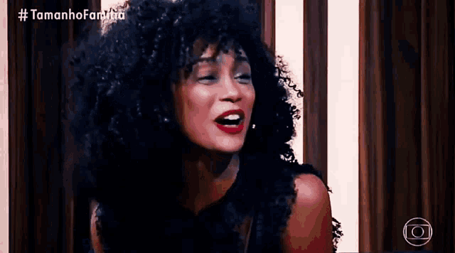 a woman with curly hair and red lipstick is sitting in front of a wooden wall and a sign that says #tamanhofamilia