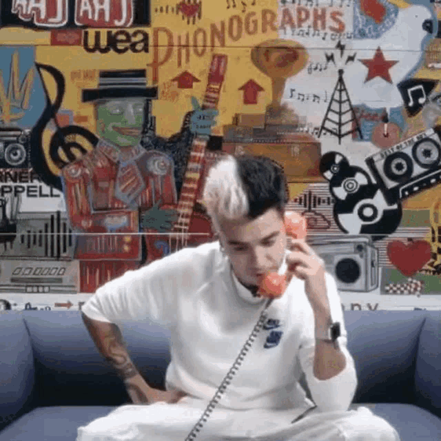 a man is sitting on a couch talking on a phone with a wall behind him that says weaphonographs