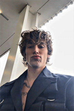 a young man with curly hair and a tattoo on his chest is smoking a cigarette