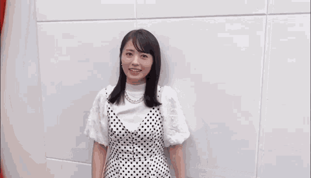 a woman in a polka dot dress is standing in front of a white wall and giving a peace sign .