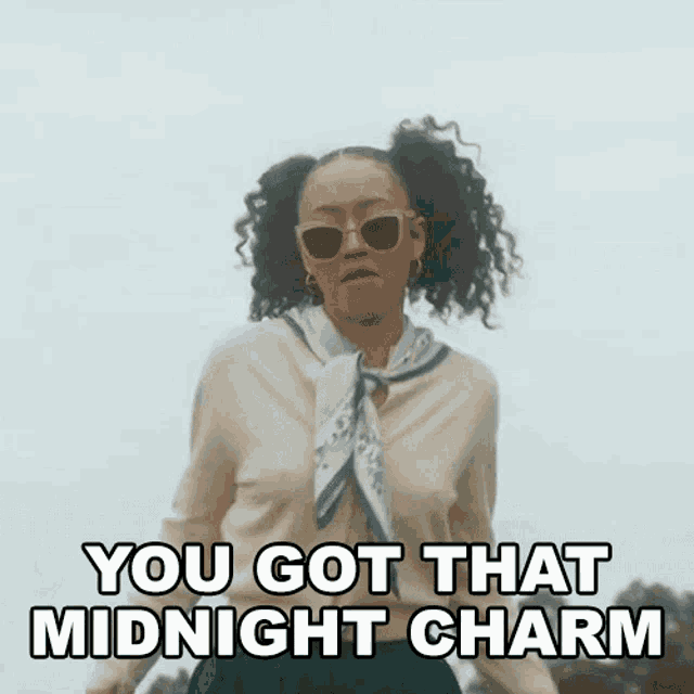 a woman wearing sunglasses and a scarf is standing in front of a sign that says `` you got that midnight charm '' .