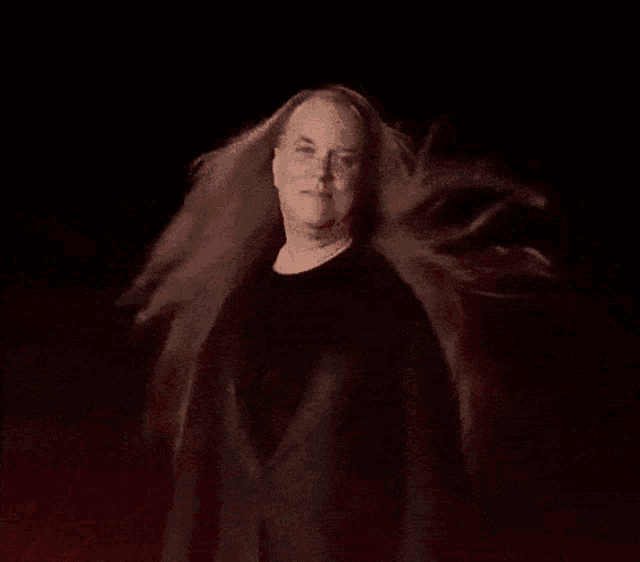 a man with long hair is standing in a dark room with a red background .