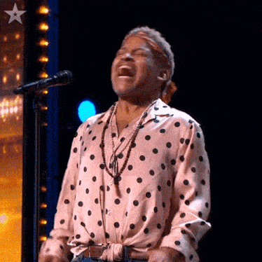 a woman in a pink polka dot shirt is singing into a microphone on a stage .