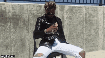 a man in a leather jacket and ripped jeans is sitting on a chair smoking a cigarette .