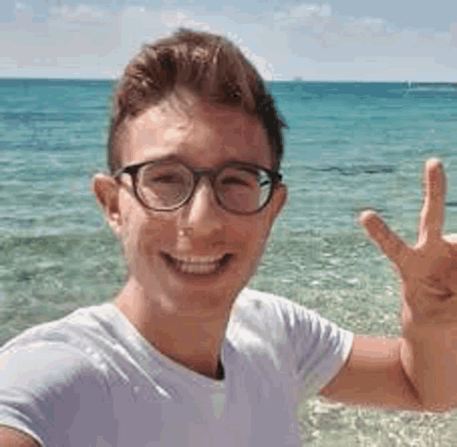a young man wearing glasses and a white shirt is taking a selfie at the beach .