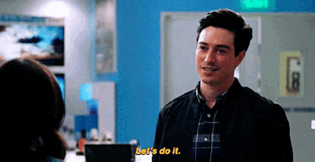 a man in a plaid shirt is talking to a woman in a room and saying let 's do it .