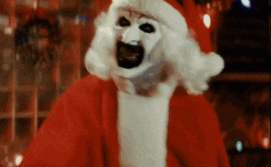 a clown dressed as santa claus is standing in front of a christmas tree and looking at the camera .