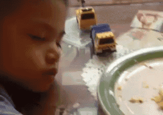 a little girl is sitting at a table with a plate of food and toy cars on it .