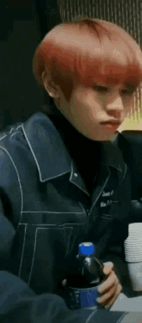a man with red hair is sitting at a table holding a bottle of soda .