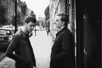 two men are standing next to each other on a sidewalk and talking to each other .