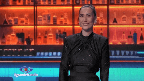a woman in a black dress is standing in front of a wall of bottles of alcohol .