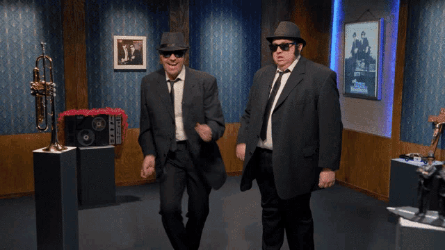 two men in suits and hats are dancing in a room with a trumpet on display