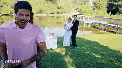 a man in a pink polo shirt is standing in front of a bride and groom holding hands .