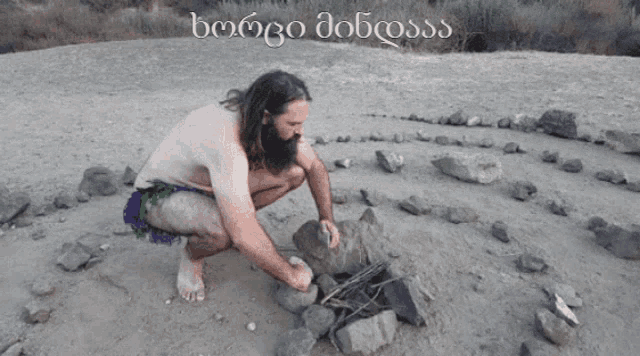 a shirtless man squatting in the sand with a foreign language on the bottom