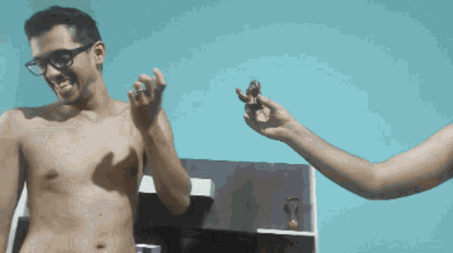 a shirtless man with glasses is smiling while a woman holds a small object