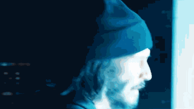 a man with a beard is wearing a blue beanie and looking at a computer screen .