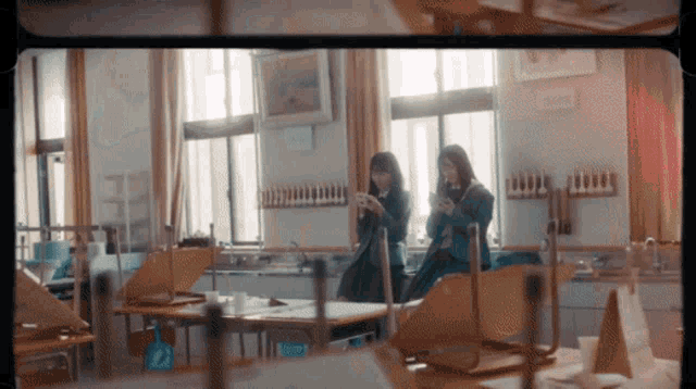 two girls are standing in an empty classroom looking at their cell phones