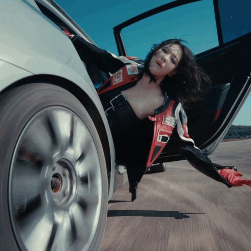 a woman in a race suit is laying on the ground next to a silver car