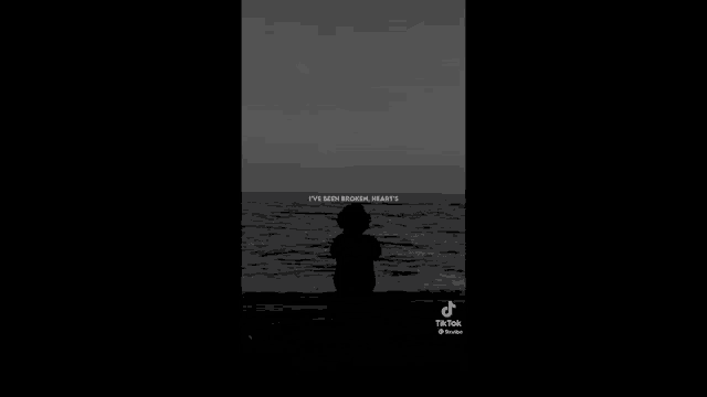 a black and white photo of a person sitting on the beach looking at the ocean .