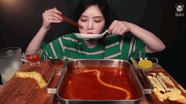 a woman in a green striped shirt is eating a hot pot with chopsticks