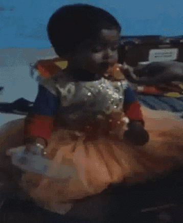 a little girl in a dress is sitting on a bed and eating an orange