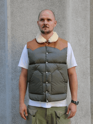 a man wearing a vest with a fur collar is standing in front of a wall