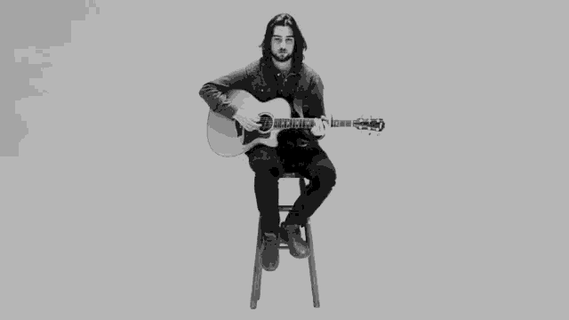 a black and white photo of a man sitting on a stool playing an acoustic guitar .