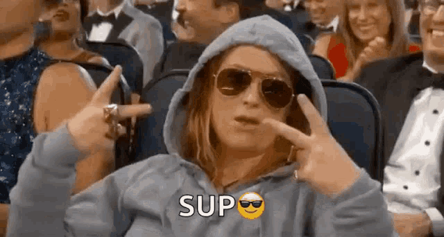 a woman wearing sunglasses and a hoodie is sitting in a crowd of people and making a peace sign .