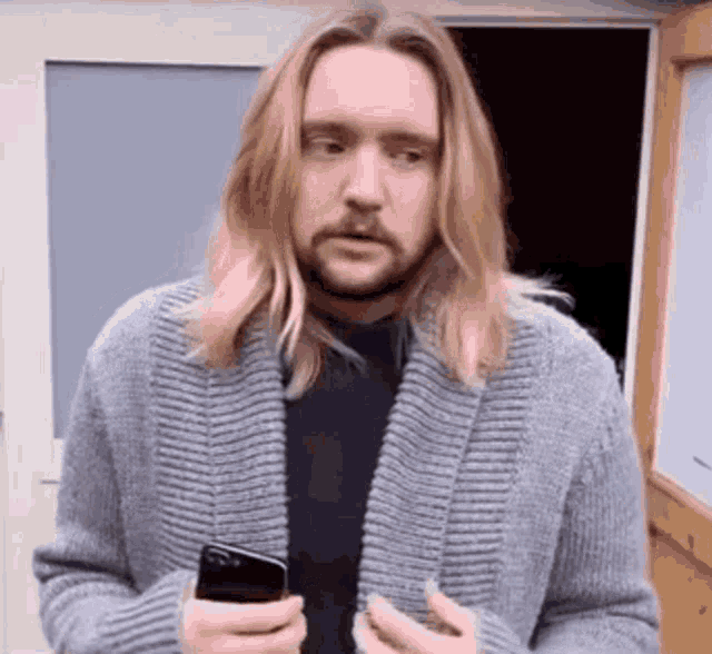 a man with long blonde hair and a beard is standing in front of a door holding a cell phone .