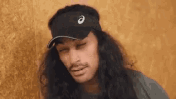 a man with long hair is wearing a black visor and a hat .