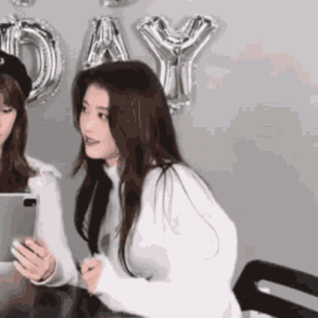 two women are sitting at a table with balloons in the background and one of them is holding a tablet .