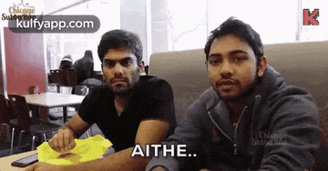 two men are sitting at a table in a restaurant and one of them is saying aithe