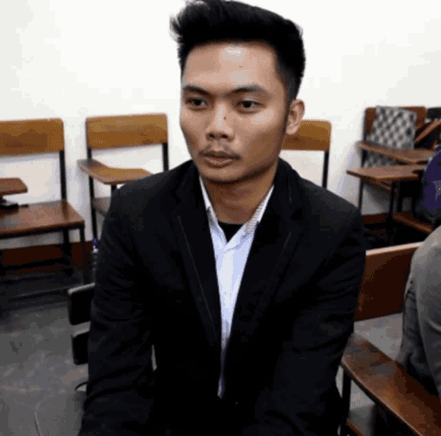 a man in a suit sits in a classroom with wooden desks