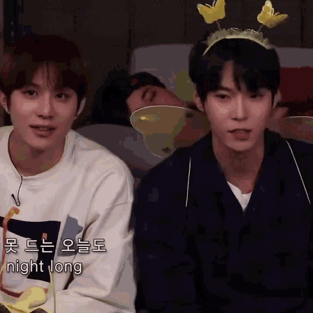 two young men are sitting next to each other on a couch . one of the men is wearing a butterfly headband .