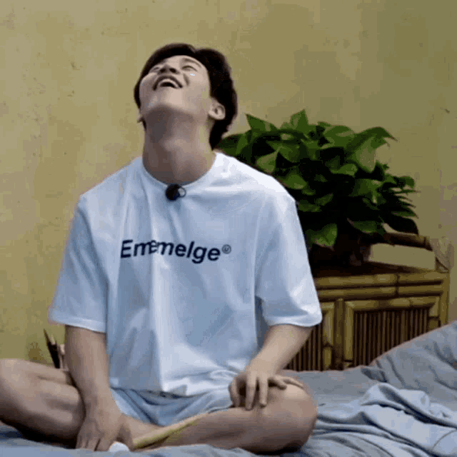 a man sitting on a bed wearing a white t-shirt that says emeelge on it