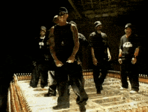 a group of men are standing in a dark room and one of them has a tattoo on his arm that says ' x2 '
