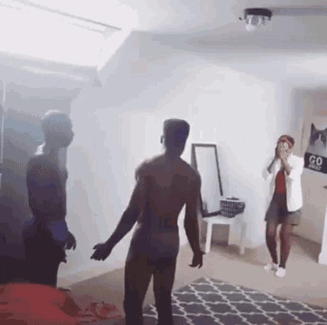 a man without a shirt is standing in a living room with a woman taking a picture of him .