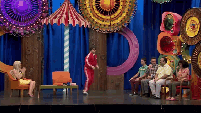 a group of people sitting in chairs on a stage with a man in a red jacket dancing