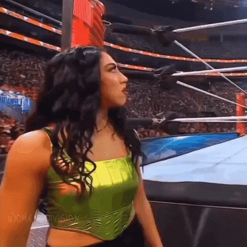 a woman is standing in a wrestling ring wearing a green top and black pants .