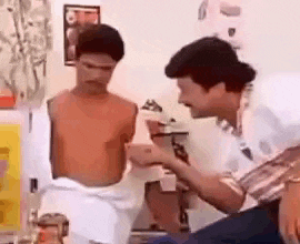 two shirtless men are standing next to each other in a room . one of the men is holding a cell phone .