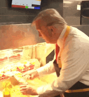 a man in a white shirt and black apron prepares food in front of a monitor that says ' a ' on it