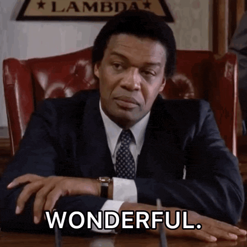 a man in a suit and tie sits at a desk with his hands on his hips and says wonderful