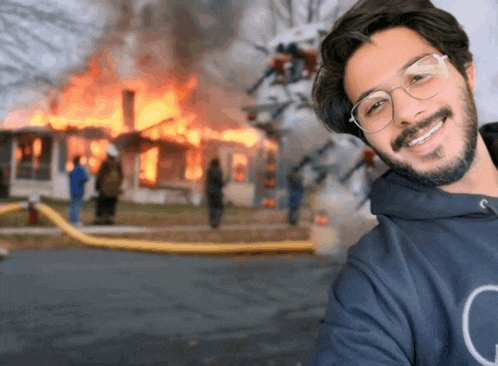 a man wearing glasses and a blue sweatshirt with the letter g on it smiles in front of a burning house