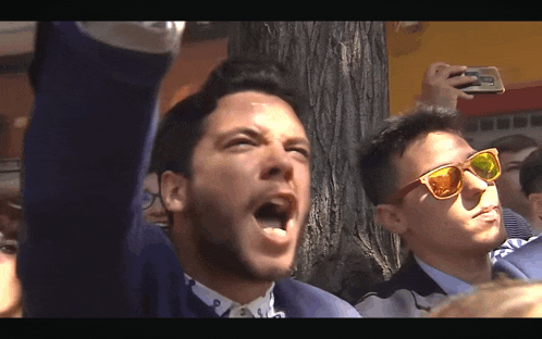 a man wearing sunglasses shouts while another man takes a picture with his phone